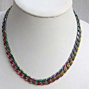 May include: A rainbow-colored chain necklace displayed on a white mannequin bust. The necklace features a thick, flat curb chain design with a metallic finish. The colors shift from red to green and blue.