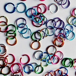 May include: A collection of small, open jump rings in a variety of vibrant colors including blue, green, pink, orange, purple, silver, and gold. Each ring features a single cut opening, allowing them to be linked together for jewelry making.