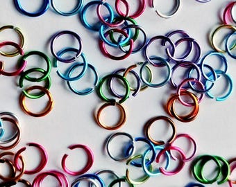 May include: A collection of small, open jump rings in a variety of vibrant colors including blue, green, pink, orange, purple, silver, and gold. Each ring features a single cut opening, allowing them to be linked together for jewelry making.