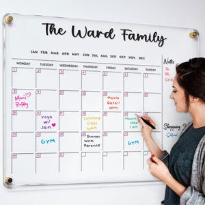 May include: A clear acrylic dry-erase calendar with the text "The Ward Family" at the top. The calendar has a grid for each month of the year, with days of the week labeled. There are handwritten notes on the calendar, including "Mom's Birthday", "Yoga w/ Jen", "Cycling Class", "Movie Date!!", "Dinner with Parents", "Mount Hike 1 am", and "Shopping Weekend - Oxford".