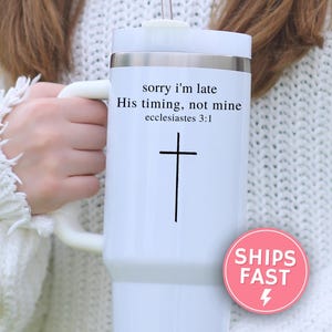 May include: White travel mug with a handle and a silver rim. The mug features the text "sorry I'm late, His timing, not mine, ecclesiastes 3:1" and a black cross. A pink "SHIPS FAST" sticker is visible.