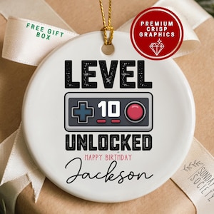 May include: A white ceramic ornament with a gold string for hanging. The ornament features the words "LEVEL 10 UNLOCKED" with a controller graphic. The name "Jackson" and "Happy Birthday" are also included. A red circle with "PREMIUM CRISP GRAPHICS" is in the upper right.