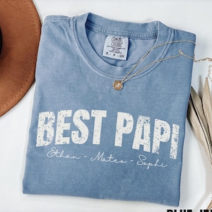 May include: A blue jean colored t-shirt with the text "BEST PAPI" in white, along with the names Ethan, Mateo, and Sophi. A gold necklace and a brown hat are also visible.