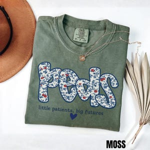 K&ouml;nnte beinhalten: Moosgr&uuml;nes T-Shirt mit dem Wort "Peds" in floralem Design. Das Shirt zeigt auch den Text "little patients, big futures" und ein kleines blaues Herz. Eine goldene Halskette und ein brauner Hut sind ebenfalls zu sehen.