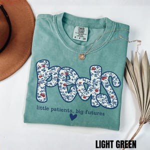 K&ouml;nnte beinhalten: Ein hellgr&uuml;nes T-Shirt mit dem Wort "Peds" in einem floralen Design. Das Shirt hat auch den Text "little patients, big futures" und ein blaues Herz. Eine silberne Halskette mit Anh&auml;nger befindet sich auf dem Shirt.