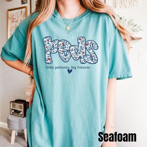 K&ouml;nnte beinhalten: T&uuml;rkisfarbenes T-Shirt mit dem Wort "Peds" in einem floralen Design. Der Text "little patients, big futures" und ein blaues Herz befinden sich unter dem Wort "Peds". Das Wort "Seafoam" steht unten rechts.