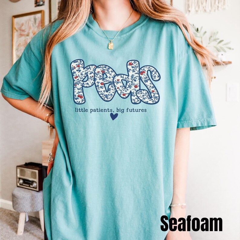 K&ouml;nnte beinhalten: T&uuml;rkisfarbenes T-Shirt mit dem Wort "Peds" in einem floralen Design. Der Text "little patients, big futures" und ein blaues Herz befinden sich unter dem Wort "Peds". Das Wort "Seafoam" steht unten rechts.