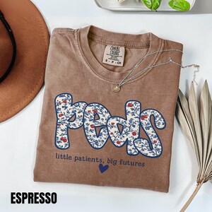 K&ouml;nnte beinhalten: Espressofarbenes T-Shirt mit dem Wort "Peds" in einem blau-wei&szlig;en Blumenmuster. Der Text "little patients, big futures" und ein blaues Herz befinden sich darunter. Eine silberne Halskette liegt auf dem Shirt.