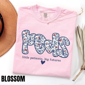 K&ouml;nnte beinhalten: Hellrosa T-Shirt mit dem Wort "Peds" in floralem Design. Das Shirt hat auch den Text "little patients, big futures" und ein blaues Herz. Das Wort "BLOSSOM" steht unten.