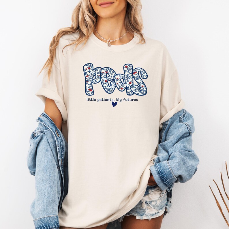 K&ouml;nnte beinhalten: Cremefarbenes T-Shirt mit dem Wort "Peds" in einem blauen Blumenmuster. Der Text "little patients, big futures" steht darunter, mit einem blauen Herzsymbol. Das Shirt hat kurze &Auml;rmel.