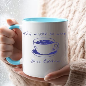 Puede incluir: Taza de cerámica blanca con interior y asa azul claro. La taza presenta un dibujo azul de una taza de café y un platillo, con el texto "This might be wine Boss Edition" en escritura azul. La taza es sostenida por una persona.