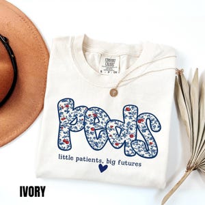 K&ouml;nnte beinhalten: Elfenbeinfarbenes Sweatshirt mit dem Wort "Peds" in einem blauen Blumenmuster. Der Text "little patients, big futures" und ein blaues Herz sind darunter. Eine goldene Halskette und ein brauner Hut sind ebenfalls im Bild.