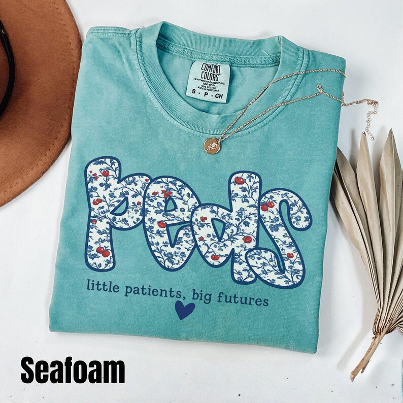 K&ouml;nnte beinhalten: T&uuml;rkisgr&uuml;nes T-Shirt mit dem Wort "peds" in einem floralen Design. Das Shirt zeigt auch den Text "little patients, big futures" und ein kleines blaues Herz. Eine goldene Halskette mit Anh&auml;nger ist &uuml;ber dem Shirt drapiert.