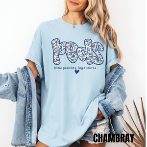 K&ouml;nnte beinhalten: Hellblaues T-Shirt mit dem Wort "Peds" in floralem Design. Der Text "little patients, big futures" und ein blaues Herz befinden sich darunter. Eine Jeansjacke ist &uuml;ber den Schultern getragen.