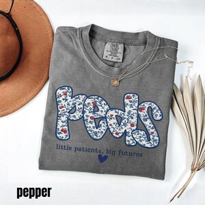 K&ouml;nnte beinhalten: Ein graues T-Shirt mit dem Wort "Peds" in einem floralen Design, mit dem Text "little patients, big futures" und einem blauen Herzen. Eine silberne Halskette mit Anh&auml;nger befindet sich auf dem Shirt.