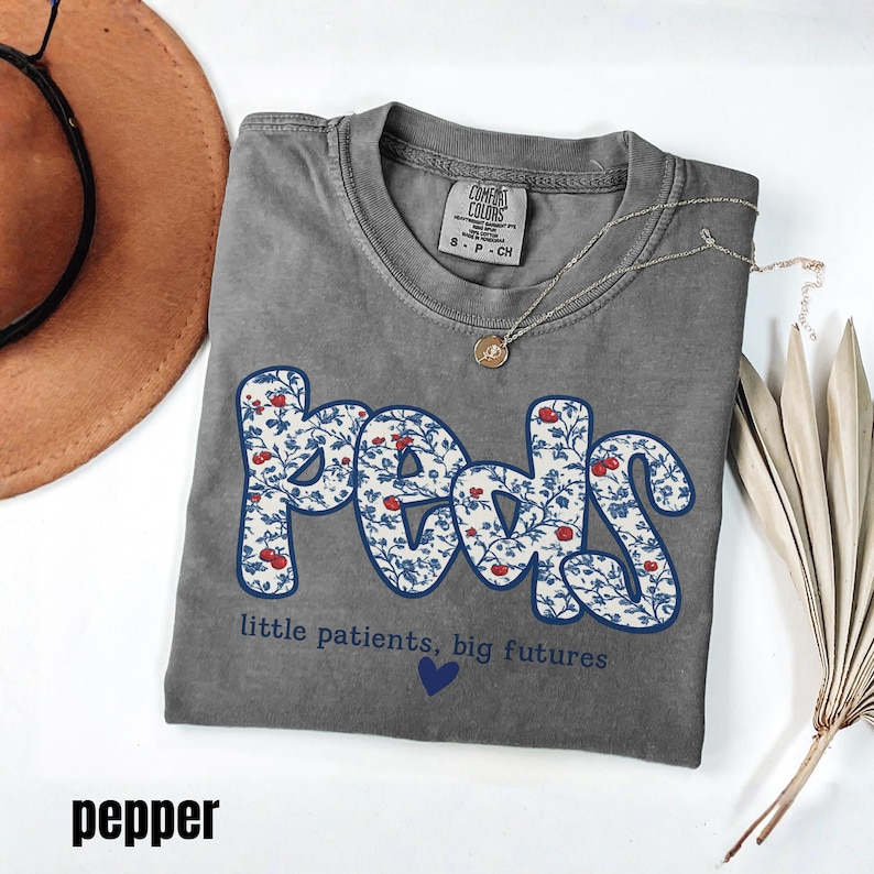 K&ouml;nnte beinhalten: Ein graues T-Shirt mit dem Wort "Peds" in einem floralen Design, mit dem Text "little patients, big futures" und einem blauen Herzen. Eine silberne Halskette mit Anh&auml;nger befindet sich auf dem Shirt.