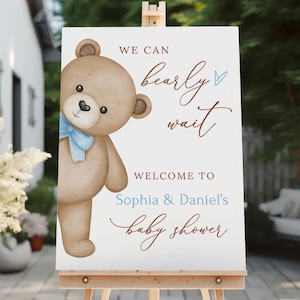 May include: A white welcome sign with a brown teddy bear peeking around the corner. The sign says "We can barely wait. Welcome to Sophia & Daniel's baby shower."