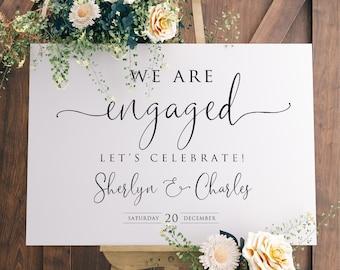 May include: White sign with black text announcing an engagement. The sign reads "We are engaged, Let's Celebrate!  Sherlyn & Charles, Saturday 20 December 2023". The sign is decorated with white and yellow flowers.