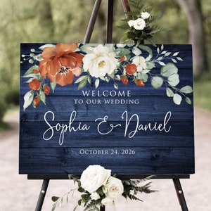 May include: A navy blue wooden sign with a white floral design and the text "Welcome to our wedding Sophia & Daniel October 24, 2026".