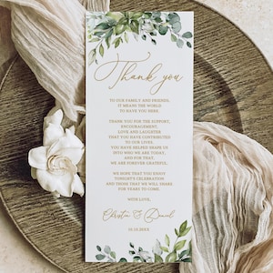 May include: A rectangular white card with the words "Thank you" in gold script, adorned with green foliage. The card includes a message of gratitude and is set on a decorative plate with a fabric overlay.