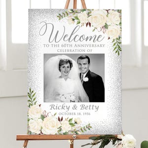 May include: A welcome sign for a 60th anniversary celebration. The sign features a black and white photo of a couple, surrounded by a floral border of white roses and greenery. The text reads "Welcome" and "Ricky & Betty, October 18, 1956."