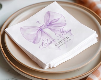 May include: White paper napkins featuring a purple watercolour bow and cross design. The text on the napkin reads "Chloe May Baptized April 20, 20XX". The napkins are stacked on a beige plate.