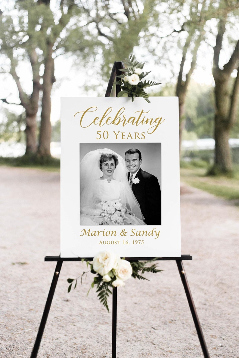 May include: White sign with gold text celebrating 50 years of marriage. The sign features a black and white photo of a couple on their wedding day and the text "Celebrating 50 Years", "Marion & Sandy", and "August 16, 1975".