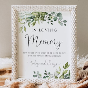 Op de afbeelding: Een ingelijst bord met de woorden "IN LOVING Memory" in elegant schrift, versierd met aquarel groen. De tekst "FOR THOSE WHO CANNOT BE HERE TODAY, BUT ARE ALWAYS IN OUR HEARTS" wordt ook weergegeven. Het bord is ingelijst in een verweerde witte lijst.