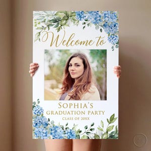 May include: A white graduation party sign featuring a photograph of a woman. The sign is adorned with blue hydrangea flowers and green foliage. Gold text reads "Welcome to" and "Sophia's Graduation Party, Class of 20XX."