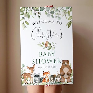 May include: Welcome to Christine's Baby Shower sign with a green leafy border and woodland animal illustrations including a deer, owl, bear, raccoon, fox, and squirrel. The date is August 29, 2026.