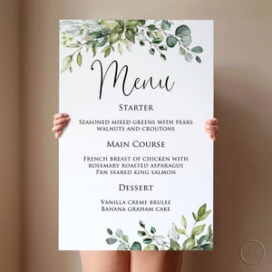 May include: A white menu board with the word "Menu" in elegant script. The menu includes starter, main course, and dessert options, with floral accents of green leaves and white flowers. The menu items are clearly listed.