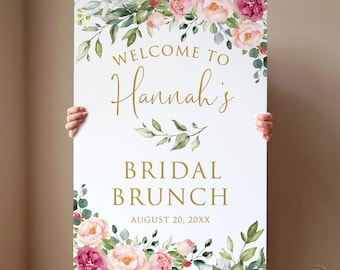 Op de afbeelding: Een wit bord met gouden tekst en bloemaccenten. Het bord leest "Welcome to Hannah's Bridal Brunch August 20, 20XX." Het bloemstuk bevat roze en rode rozen met groene bladeren, die de boven- en onderkant van het bord omlijsten.
