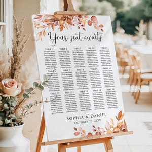 May include: A white wedding seating chart sign on a wooden easel featuring the text Your seat awaits, Sophia & Daniel, October 20, 20XX, and fifteen tables with guest names. The sign is decorated with watercolor autumn leaves in orange and brown.
