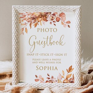 May include: A framed sign with the words "Photo Guestbook" in gold script. The sign includes the text "Snap it, Stick it, Sign it" and "Please leave a photo and well wishes for Sophia." The frame is white with a textured pattern, and the sign is decorated with watercolor floral elements.