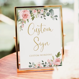 May include: A gold-framed sign with the words "Custom Sign" in elegant gold script. The sign is decorated with watercolor-style pink roses and greenery. The bottom of the sign reads "Custom Wording".