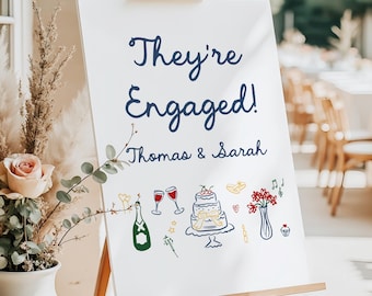 May include: White sign on a wooden easel with the text "They're Engaged!" in navy blue script. Below, the names "Thomas & Sarah" are written. The sign is decorated with colorful illustrations of champagne, cake, and flowers.