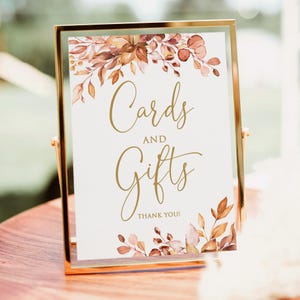 May include: A gold-framed sign with the words "Cards and Gifts" in elegant gold script. The sign is decorated with watercolor-style floral elements in shades of brown, orange, and beige. The words "Thank You!" are at the bottom.