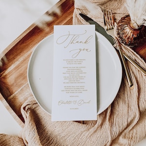 May include: A white card with gold lettering that says "Thank you" and includes a message of gratitude to family and friends. The card is on a white plate with a brown napkin and a gold vase with dried flowers.