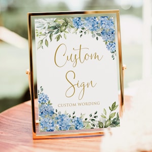 May include: A gold-framed sign featuring the words "Custom Sign" in gold script. The sign is adorned with watercolour blue hydrangea and green foliage. The words "Custom Wording" are printed at the bottom.