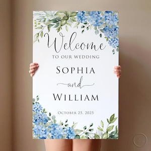 May include: A white wedding welcome sign with the text "Welcome to our wedding" above the names "Sophia and William" and the date "October 25, 2025". The sign is decorated with blue hydrangea flowers and green leaves.