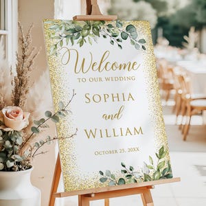 May include: A white wedding welcome sign with gold glitter accents and green foliage. The sign reads "Welcome to our Wedding" followed by the names "Sophia and William" and the date "October 25, 20XX". The sign is displayed on a wooden easel.