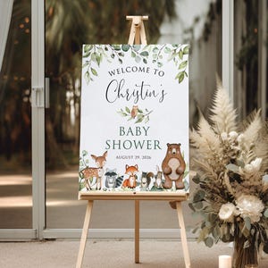 May include: Welcome to Christin's Baby Shower sign with a woodland theme. The sign features a brown bear, a fox, a deer, a raccoon, and a squirrel. The sign is on a wooden easel and has a green leafy border.
