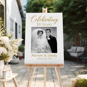 May include: White sign with gold text celebrating 50 years of marriage. A black and white photo of a couple on their wedding day is in the center of the sign. The text reads "Celebrating 50 Years" and "Marion & Sandy August 16, 1975".