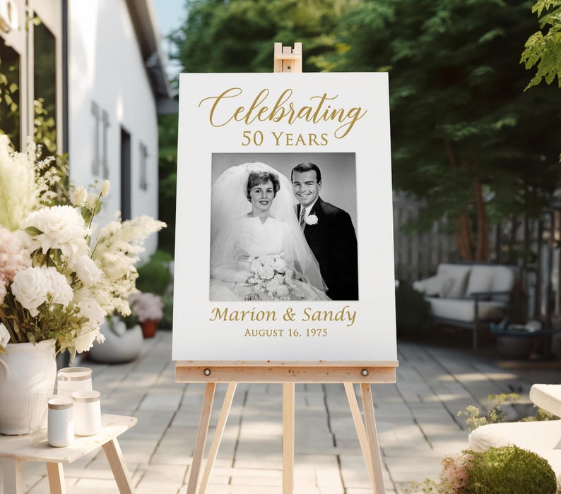May include: White sign with gold text celebrating 50 years of marriage. A black and white photo of a couple on their wedding day is in the center of the sign. The text reads "Celebrating 50 Years" and "Marion & Sandy August 16, 1975".