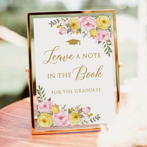 May include: A gold framed sign with the text "Leave a Note in the Book for the Graduate." The sign is decorated with watercolor-style pink and yellow flowers and greenery. A gold graduation cap graphic is above the text.