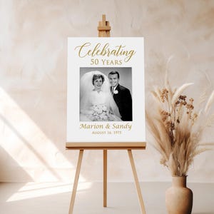 May include: A white sign with gold text celebrating 50 years of marriage. The sign features a black and white photo of a couple in their wedding attire. The text reads "Celebrating 50 Years" and "Marion & Sandy August 16, 1975".