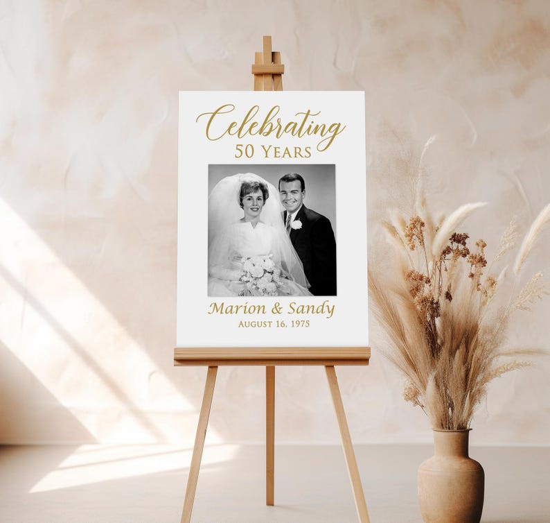 May include: A white sign with gold text celebrating 50 years of marriage. The sign features a black and white photo of a couple in their wedding attire. The text reads "Celebrating 50 Years" and "Marion & Sandy August 16, 1975".