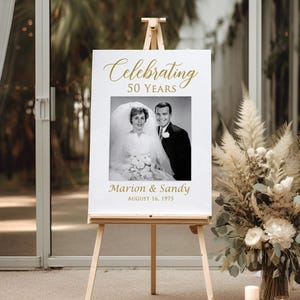 May include: A white sign with gold text celebrating 50 years of marriage. The sign features a black and white photo of a couple in their wedding attire. The text reads "Celebrating 50 Years" and "Marion & Sandy August 16, 1975".