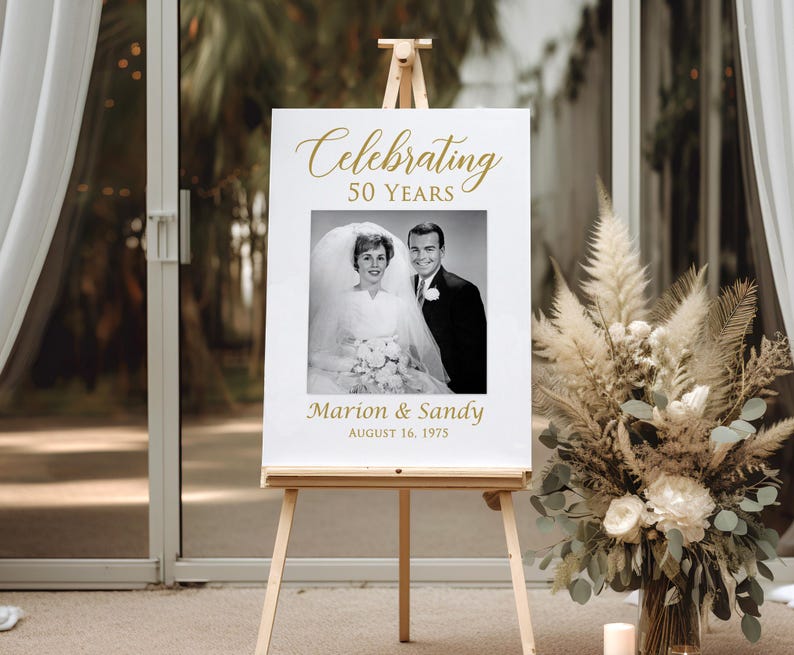 May include: A white sign with gold text celebrating 50 years of marriage. The sign features a black and white photo of a couple in their wedding attire. The text reads "Celebrating 50 Years" and "Marion & Sandy August 16, 1975".