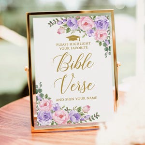 以下が含まれることがあります： 「Bible Verse」の文字がエレガントな筆記体で書かれた、金色のフレームのサイン。ピンクと紫の水彩画の花で飾られています。サインには「Please Highlight Your Favorite」と「And Sign Your Name」の文字も含まれています。